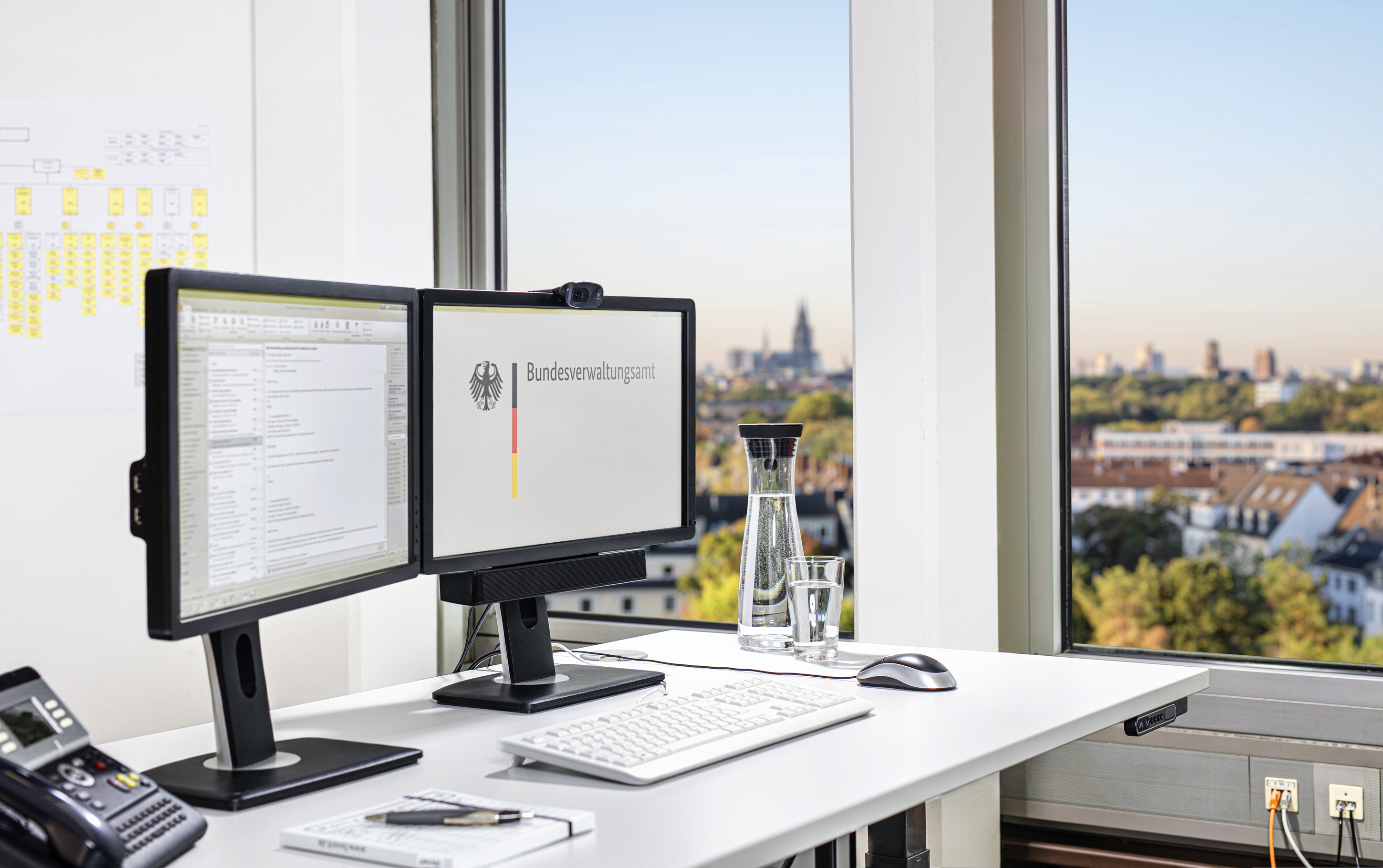 Schreibtisch mit zwei Bildschirmen, einer Tastatur und Computer-Maus, einem Telefon sowie ein Notizblock mit Stift. Vom Schreibtisch aus blickt man aus dem Fenster mit dem Kölner Dom im leicht verschwommenen Hintergrund.